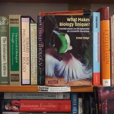 Ernst Mayr's What Makes Biology Unique? facing out on a bookstore shelf, its cover showing a bird with an iridescent green head and long white plumes, between the spines of other books by Mayr and on evolution, above a handwritten shelf label reading Science & Nature (Biology, Botany, Zoology, Evolution)