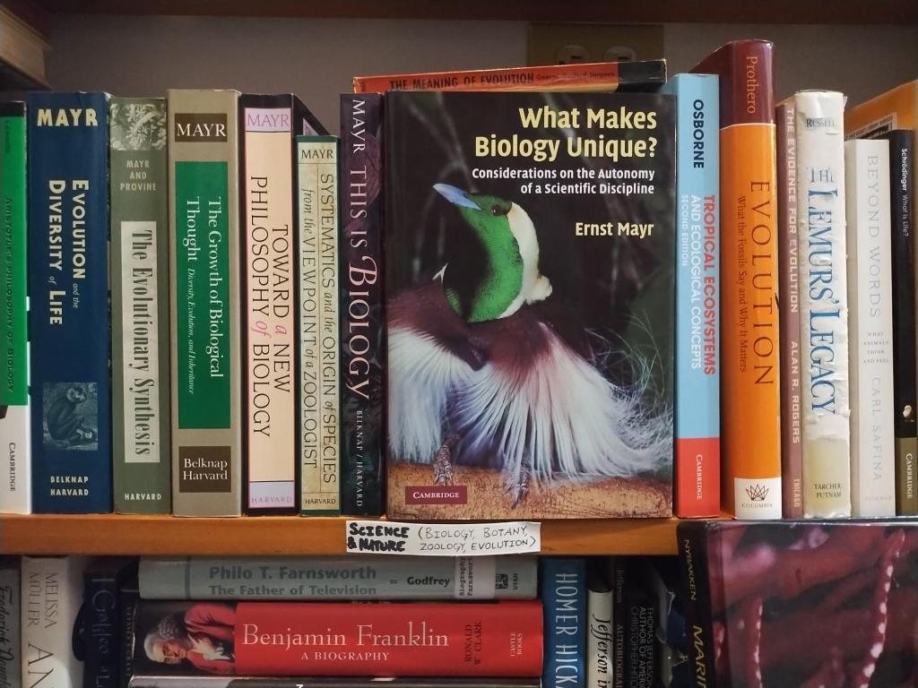 Ernst Mayr's What Makes Biology Unique? facing out on a bookstore shelf, its cover showing a bird with an iridescent green head and long white plumes, between the spines of other books by Mayr and on evolution, above a handwritten shelf label reading Science & Nature (Biology, Botany, Zoology, Evolution)