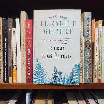 The Spanish edition of Elizabeth Gilbert's The Signature of All Things, La firma de todas las cosas, its white cover with blue ferns facing out on a wooden bookshelf among Spanish-language books on Ecuador and Galápagos