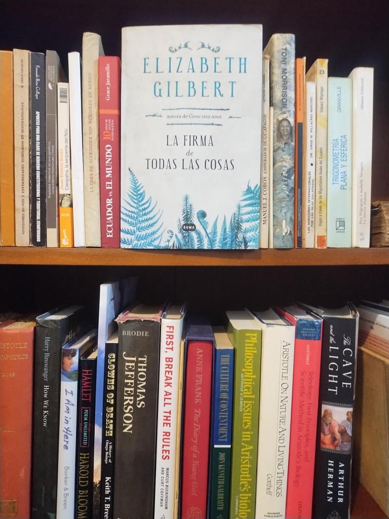 The Spanish edition of Elizabeth Gilbert's The Signature of All Things, La firma de todas las cosas, facing out on a bookshelf, with a shelf of books on Aristotle and philosophy below it